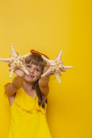 Téléchargez les photos : Portrait d'une belle petite fille tenant des étoiles de mer, rêvant de vacances d'été à la plage, isolée sur fond jaune - en image libre de droit