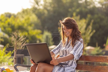 Téléchargez les photos : Femme pigiste assise sur un banc sur une terrasse dans la cour arrière, travaillant sur un ordinateur portable, buvant du café et profitant d'une journée ensoleillée à l'extérieur ; femme télétravail - en image libre de droit