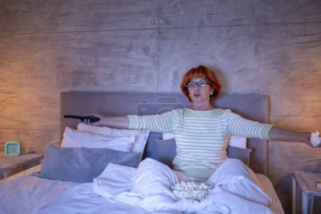 Foto de Hermosa anciana con pijamas sentada en la cama, comiendo palomitas de maíz y viendo películas de miedo en la televisión, molesta y tensa - Imagen libre de derechos