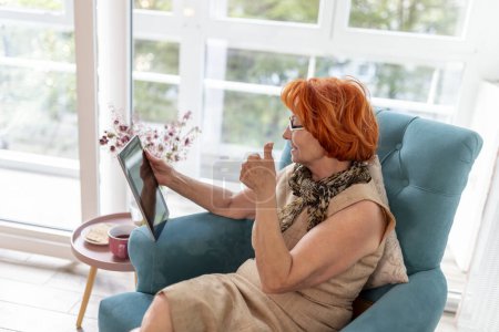 Photo for Elderly woman sitting in an armchair, relaxing at home and using a tablet computer; senior woman having a video call on tablet computer - Royalty Free Image