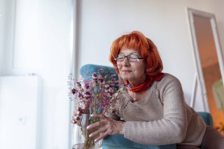 Photo for Senior woman sitting in an armchair, arranging a flower bouquet into a vase and relaxing at home - Royalty Free Image