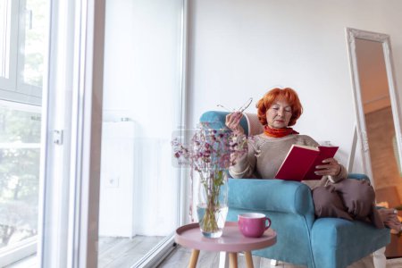 Téléchargez les photos : Belle femme âgée assise dans un fauteuil près de la fenêtre, lisant un livre et se relaxant à la maison - en image libre de droit