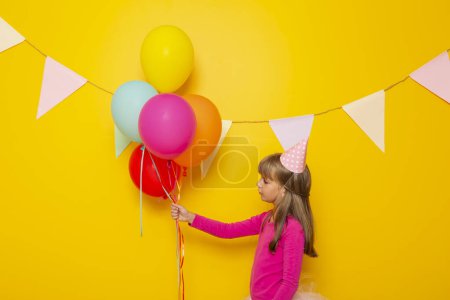 Photo for Beautiful little girl having fun celebrating her birthday, holding bunch of colorful balloons, isolated on yellow colored background - Royalty Free Image