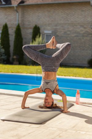 Téléchargez les photos : Belle jeune femme active faisant la pose de la tête de yoga, pratiquant le yoga à l'extérieur près de la piscine par une journée d'été ensoleillée - en image libre de droit