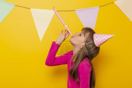 Foto de Hermosa niña divirtiéndose celebrando su cumpleaños, soplando silbato de fiesta, aislado sobre fondo de color amarillo - Imagen libre de derechos