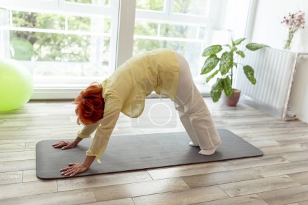 Téléchargez les photos : Femme âgée active faisant de l'entraînement de yoga à la maison, tenant vers le bas face à la pose de yoga pour chien - mode de vie sain et actif des personnes âgées concept - en image libre de droit
