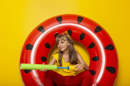 Téléchargez les photos : Petite fille jouant dans un anneau de natation gonflable de pastèque, se préparant pour des vacances d'été à la plage, isolée sur fond jaune - en image libre de droit