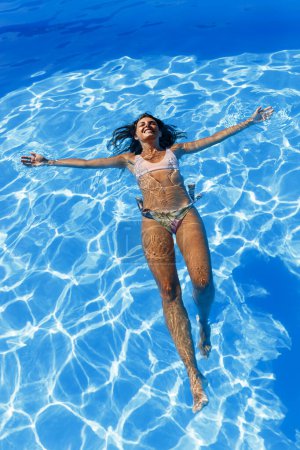 Photo for High angle view of beautiful young woman wearing swimsuit floating on water in swimming pool relaxing while on summer vacation - Royalty Free Image
