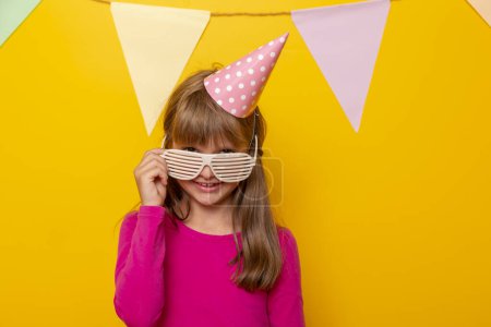 Photo for Portrait of a beautiful little girl wearing party glasses and birthday hat, having fun celebrating her birthday, isolated on yellow colored background - Royalty Free Image