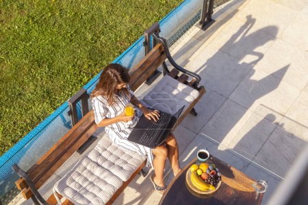 Téléchargez les photos : Vue grand angle de la jeune femme assise sur la terrasse dans la cour arrière de sa maison, à l'aide d'un ordinateur portable tout en télécommutant - en image libre de droit