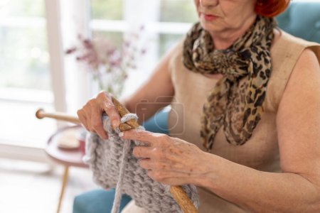Photo for Senior woman enjoying her leisure time at home, sitting in an armchair, knitting a scarf - Royalty Free Image