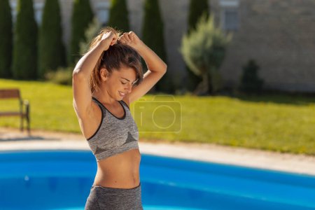 Téléchargez les photos : Fit jeune femme se préparer pour une séance d'entraînement en plein air près de la piscine, attachant ses cheveux dans une queue de cheval - en image libre de droit