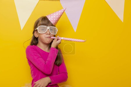 Foto de Hermosa niña divirtiéndose celebrando su cumpleaños, usando gafas de fiesta y soplando silbato de fiesta, aislado sobre fondo de color amarillo - Imagen libre de derechos