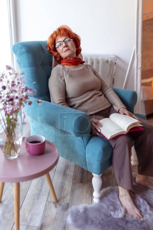 Téléchargez les photos : Femme âgée dormant dans un fauteuil avec un livre sur les genoux, faisant une sieste l'après-midi et se relaxant à la maison - en image libre de droit