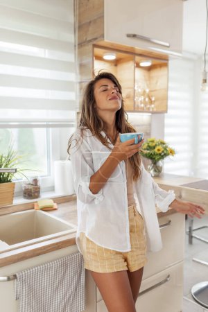 Téléchargez les photos : Belle jeune femme debout dans la cuisine, boire une tasse de café chaud, se détendre à la maison le matin - en image libre de droit