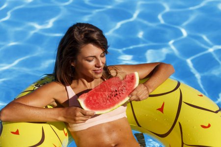Téléchargez les photos : Attrayant jeune femme en maillot de bain manger tranche de pastèque froide tout en flottant sur le radeau de la piscine, profiter d'une journée d'été ensoleillée à la piscine - en image libre de droit