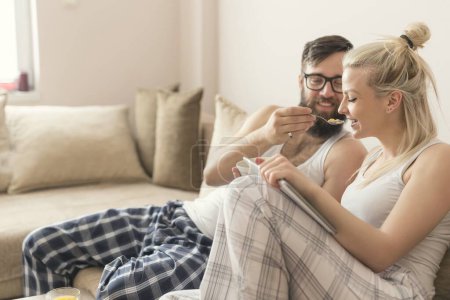 Téléchargez les photos : Couple amoureux assis sur le canapé dans le salon, en pyjama après s'être levé le matin, profiter de la matinée et prendre le petit déjeuner - en image libre de droit
