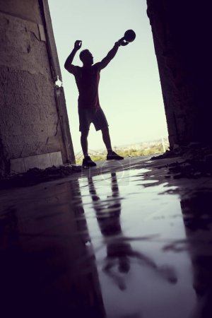 Foto de Musculoso, atlético, atleta joven haciendo ejercicio en un edificio en ruinas junto a un charco de agua. Entrenamiento Crossfit con pesas y levantamiento de pesas - Imagen libre de derechos