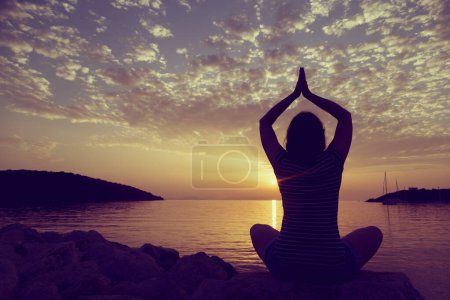 Téléchargez les photos : Silhouette d'une jeune femme pratiquant le yoga sur un rivage rocheux dans la baie de Syvota, Grèce. Ciel nuageux, mer Ionienne et un beau coucher de soleil en arrière-plan. - en image libre de droit