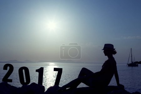 Téléchargez les photos : Jeune femme assise sur le rivage rocheux à côté d'un panneau 2017, regardant un beau coucher de soleil au port de Syvota, Grèce - en image libre de droit