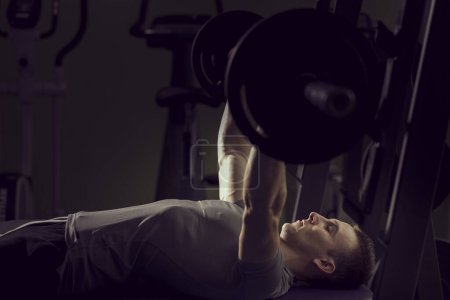 Photo for Young muscular built man lying on the bench and lifting a barbell at the gym - Royalty Free Image