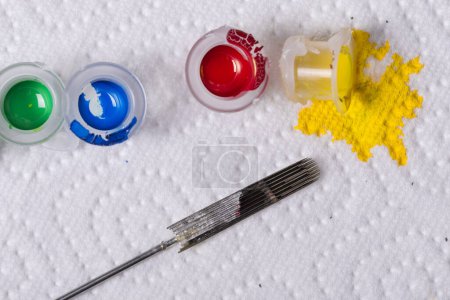 Foto de Detalle de primer plano de tintas de colores vertidas en pequeñas tazas y agujas de tatuaje aisladas sobre fondo blanco - Imagen libre de derechos