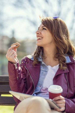 Téléchargez les photos : Jeune belle brune assise sur un banc en bois dans un parc, buvant du café, mangeant un cookie, surfant sur Internet sur une tablette et profitant d'une journée ensoleillée - en image libre de droit
