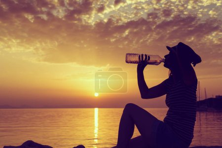 Foto de Mujer joven sentada en la costa rocosa, bebiendo agua y disfrutando de una hermosa puesta de sol en la bahía de Syvota, Grecia - Imagen libre de derechos