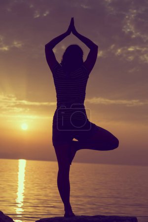 Photo for Silhouette of a young woman practicing yoga on a rocky shore in Syvota bay, Greece. Cloudy sky, Ionian sea and a beautiful sunset in the background. - Royalty Free Image