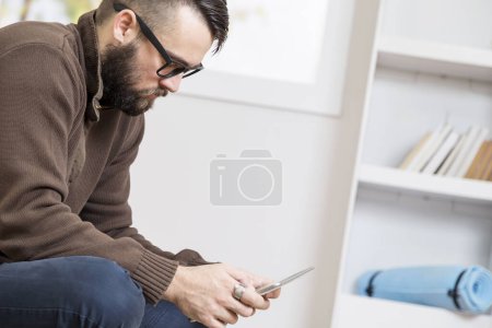 Téléchargez les photos : Homme barbu assis sur le sol, tenant une tablette et surfant sur le net - en image libre de droit