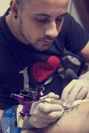 Téléchargez les photos : Tatoueur masculin tenant un pistolet de tatouage, montrant un processus de fabrication de tatouages sur le bras d'un mannequin tatoué masculin. Focus sur les yeux du tatoueur - en image libre de droit