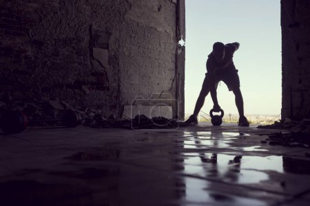 Photo for Muscular, athletic built, young athlete working out with a kettlebell in a ruin building next to a puddle of water - Royalty Free Image