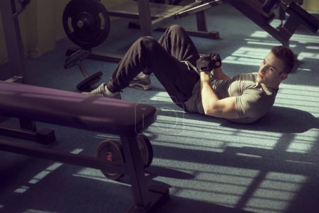 Téléchargez les photos : Jeune homme musclé attrayant travaillant dans une salle de gym, faisant des redressements assis sur le sol à côté d'un appareil d'haltérophilie - en image libre de droit