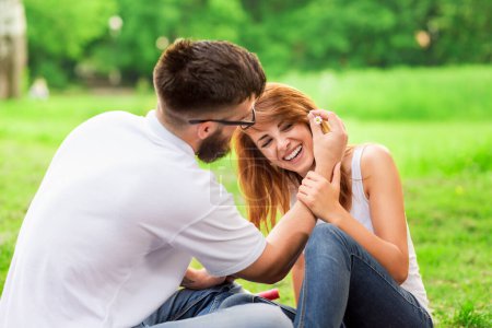 Téléchargez les photos : Couple assis sur une couverture de pique-nique dans un parc. Guy plaçant doucement des fleurs dans les cheveux de la fille - en image libre de droit