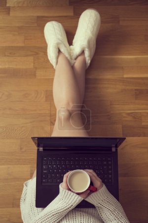 Téléchargez les photos : Vue de dessus d'une jeune femme assise sur le sol du salon, appuyée contre un canapé, buvant du café et utilisant un ordinateur portable - en image libre de droit