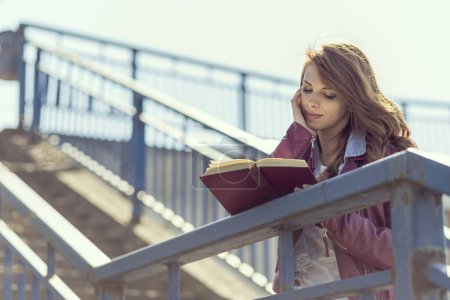 Photo for Girl standing on the stairs next to a fence and reading a book - Royalty Free Image