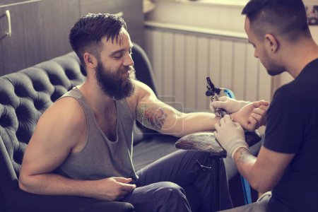 Photo for Male tattoo artist holding a tattoo gun, showing a process of making tattoos on a male tattooed model's arm. - Royalty Free Image