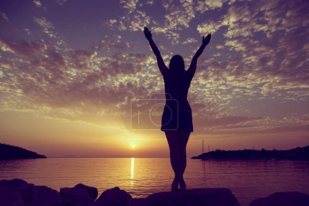 Téléchargez les photos : Jeune femme debout sur une rive rocheuse de la baie de Syvota, en Grèce, regardant le coucher du soleil et jouissant de la beauté et de la liberté - en image libre de droit