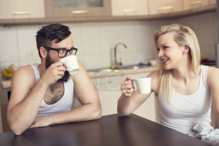 Photo for Young couple in love sitting at the table in the kitchen, drinking morning coffee. Focus on woman - Royalty Free Image