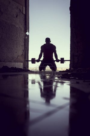 Foto de Musculoso, atlético, atleta joven haciendo ejercicio en un edificio en ruinas junto a un charco de agua. Entrenamiento Crossfit con pesas y levantamiento de pesas - Imagen libre de derechos