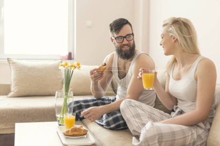 Photo for Couple in love sitting on the couch in the living room, wearing pajamas after getting up in the morning, enjoying the morning and having breakfast - Royalty Free Image
