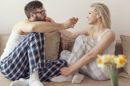 Téléchargez les photos : Couple amoureux assis sur le canapé dans le salon, portant un pyjama après s'être levé le matin, profitant de la matinée et se nourrissant mutuellement de croissants au chocolat - en image libre de droit