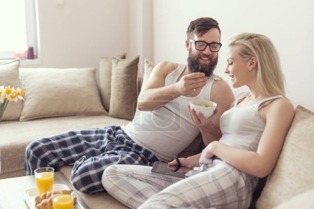 Téléchargez les photos : Couple amoureux assis sur le canapé dans le salon, portant un pyjama après s'être levé le matin, profiter de la matinée et prendre le petit déjeuner ensemble. Concentrez-vous sur le gars - en image libre de droit