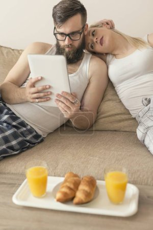 Foto de Pareja enamorada sentada en el sofá de la sala de estar, vistiendo pijamas después de levantarse por la mañana, leyendo noticias en una computadora y desayunando - Imagen libre de derechos