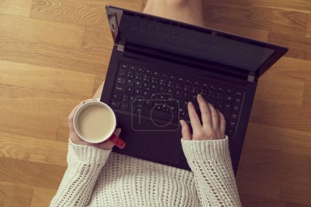 Photo for Top view of a young woman sitting on the living room floor, leaning against a couch, drinking coffee and using a laptop computer - Royalty Free Image