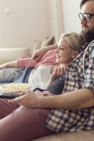 Photo for Young couple relaxing in their apartment, lying on the couch, watching a movie and eating popcorn. Focus on the girl - Royalty Free Image