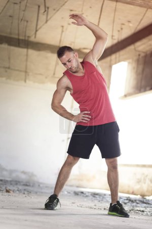Téléchargez les photos : Musclé, athlétique, jeune homme qui s'étire dans un bâtiment en ruine avant l'entraînement - en image libre de droit