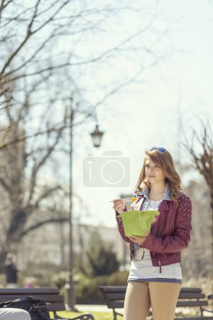 Téléchargez les photos : Jeune, belle brune assise sur un banc en bois dans un parc par une journée ensoleillée à manger de la nourriture chinoise - en image libre de droit