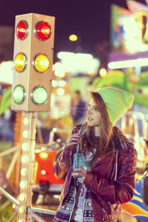 Foto de Hermosa chica urbana joven de pie junto a un semáforo en un parque de atracciones, beber bebida energética - Imagen libre de derechos