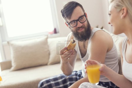 Téléchargez les photos : Couple amoureux assis sur le canapé dans le salon, en pyjama après s'être levé le matin, profiter de la matinée et prendre le petit déjeuner ensemble - en image libre de droit